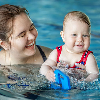 A young swimmer with a blue and white swim cap and sunglasses is performing a freestyle stroke in a bright blue swimming pool, splashing water around.